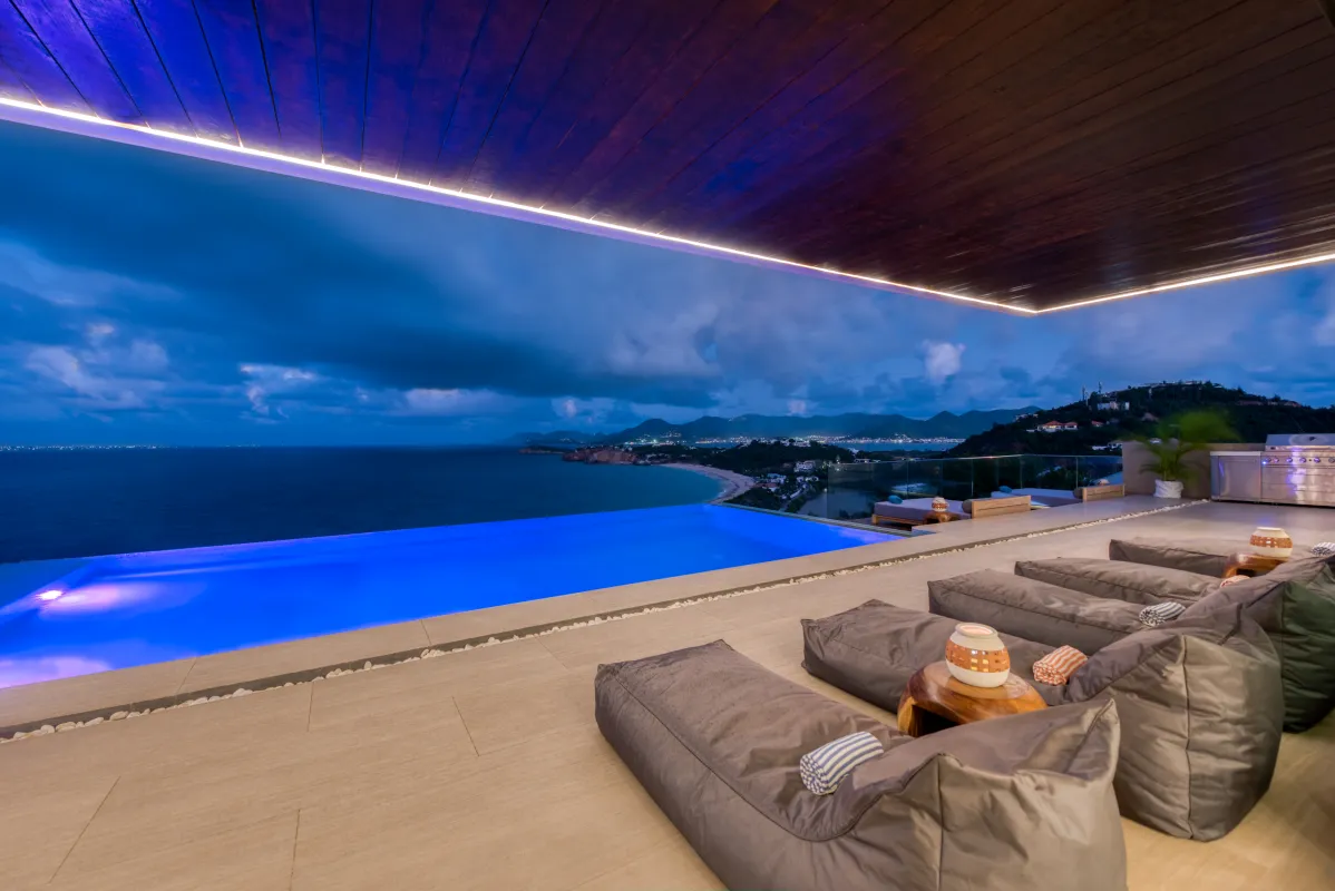 Villa Amandara: Covered terrace with lounge cushions beside an illuminated infinity pool overlooking the ocean at dusk