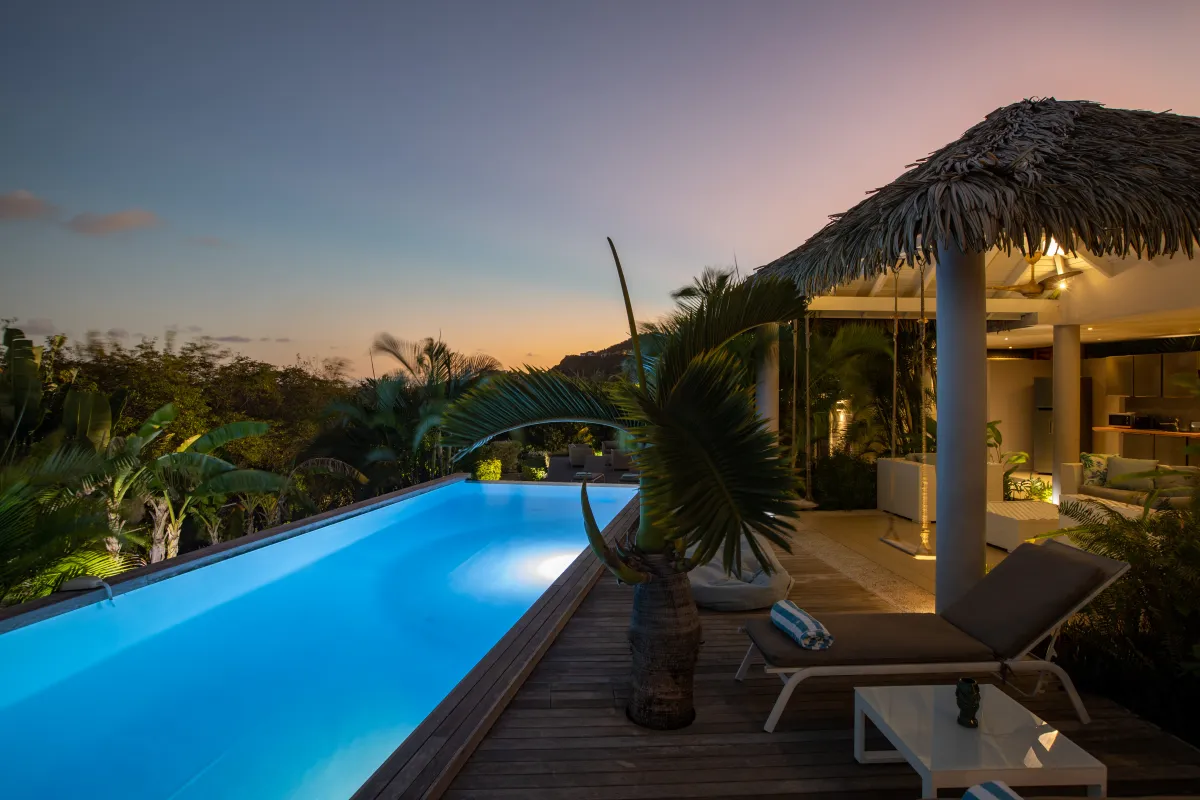 Villa Bahia Blue: Illuminated swimming pool and wooden deck with sun loungers beside a thatched pavilion at dusk