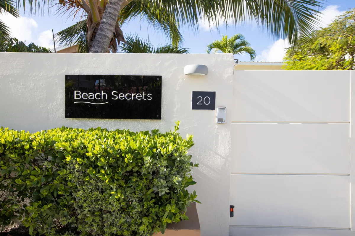 Villa Beach Secrets: White entrance wall with gate, address plaque, and green hedge under palm trees