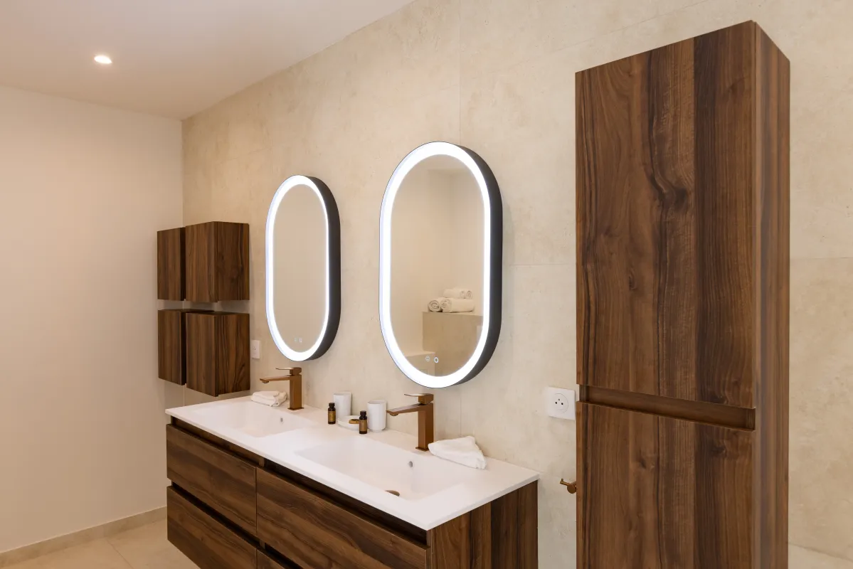 Villa Greystone: Double vanity with wood cabinetry and illuminated oval mirrors mounted on tiled wall