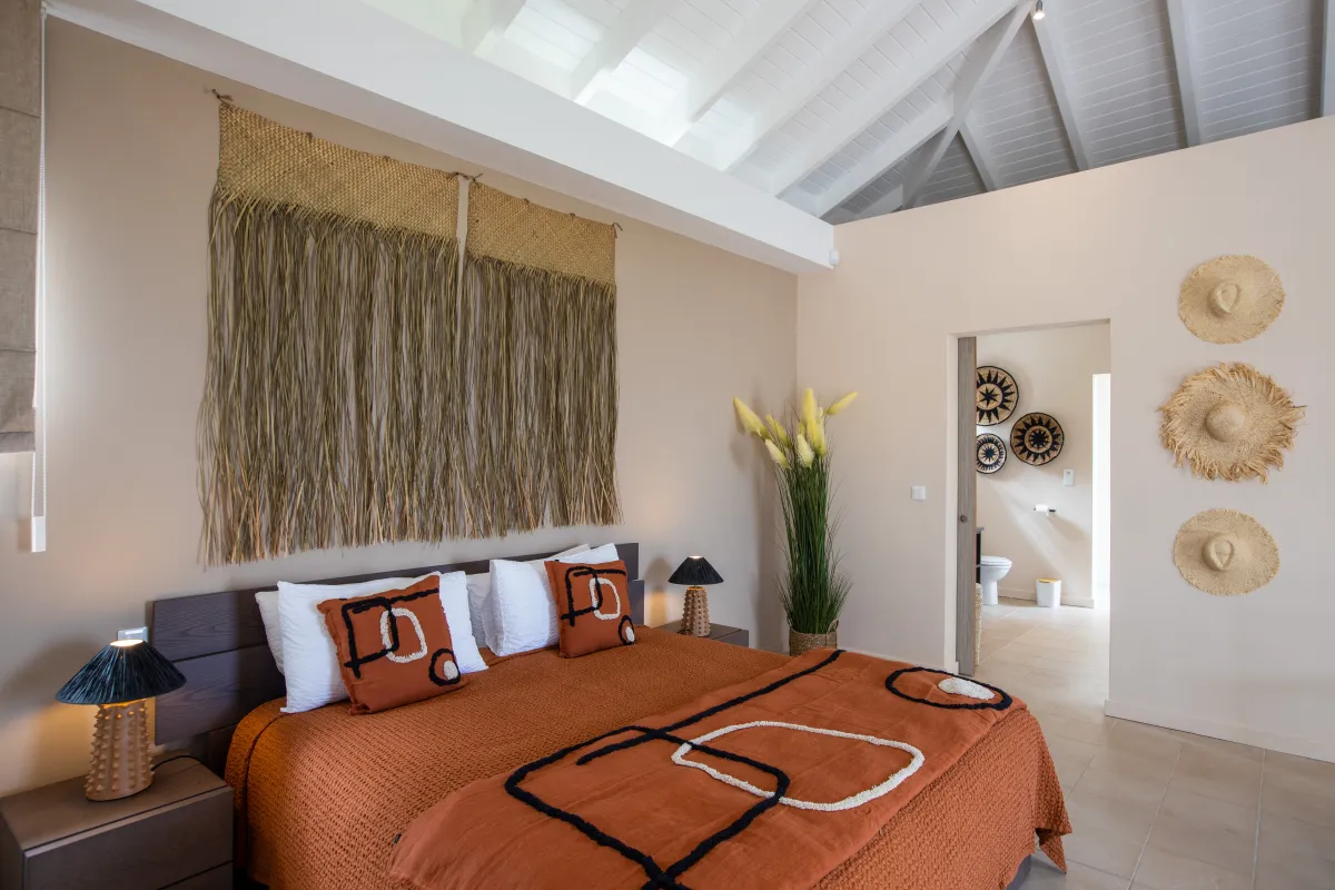 Villa Karukera: Bedroom with terracotta bedding, woven wall hanging, high ceilings, and ensuite bathroom doorway