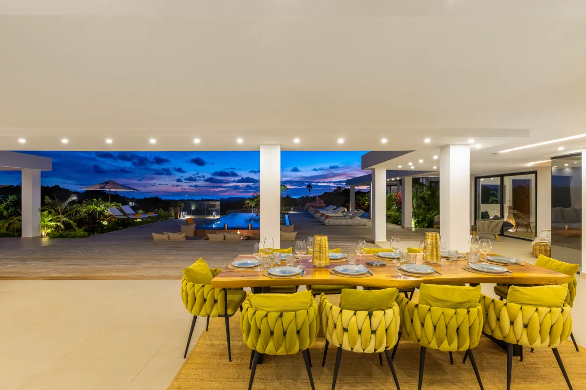 Villa Nuit Jade: Open-air dining table with modern yellow chairs overlooking a pool deck at twilight