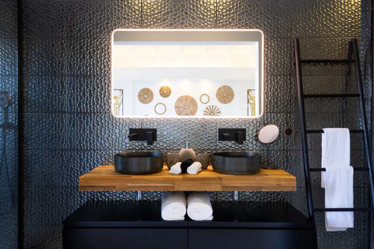 Villa Nuit Jade: Bathroom vanity with two vessel sinks, an illuminated mirror, and textured dark wall tiles